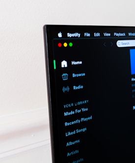 A computer screen displays the Spotify application, showing menu options such as Home, Browse, Radio, and Your Library, which includes Made For You, Recently Played, and Liked Songs. The application interface is sleek, with a dark background and white text, alongside green, yellow, and red window control buttons at the top left.