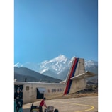 A small airplane is positioned on a runway with towering snow-capped mountains in the background, under a clear blue sky. Two people are visible; one is working near the aircraft while the other person is seated inside.