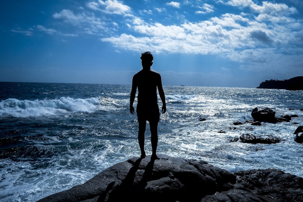 homme en veste noire debout sur un rocher près de la mer pendant la journée