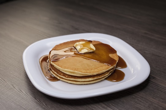 A stack of three golden-brown pancakes is topped with a melting pat of butter and generously drizzled with maple syrup. The pancakes are neatly arranged on a white square plate, which contrasts with the dark wooden surface of the table beneath.