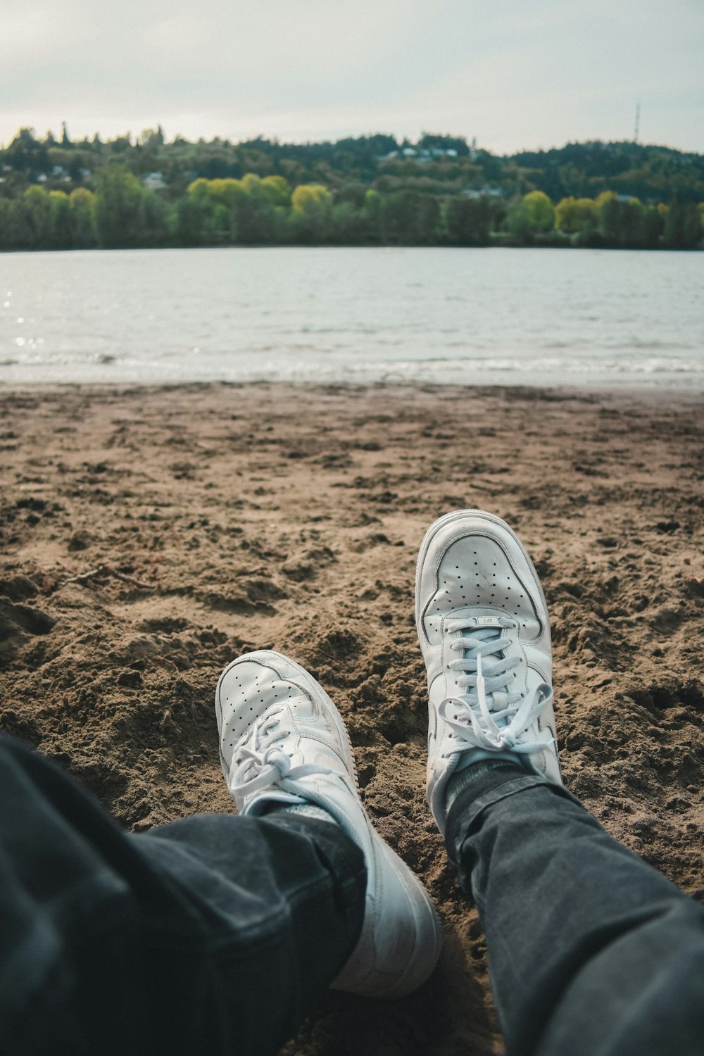 person in blue denim jeans and white sneakers sitting on brown sand near body of water
