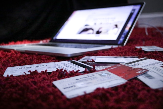 A slightly blurred laptop is open and resting on a red carpet. In the foreground, several credit cards and receipts are scattered across the carpet, suggesting a scene involving online shopping or financial transactions.