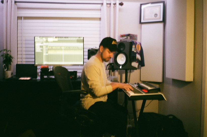 A person is sitting at a keyboard in a home recording studio. There are audio speakers, a computer monitor displaying music production software, and various equipment around the room. Certificates are framed on the wall, and a plant is on a shelf.