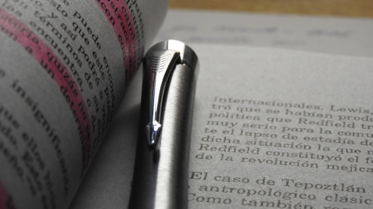 A close-up view of an open book with text highlighted in pink. A silver pen is resting against the pages, emphasizing the highlighted section.