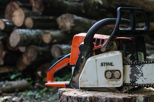 A chainsaw resting on a tree stump with stacks of cut logs in the background. The chainsaw is prominently orange and white with a logo visible on the casing. The focus is sharp on the tool, while the background appears slightly blurred, highlighting the depth of the scene.