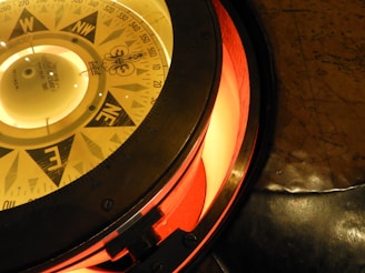 An antique nautical compass with detailed directional markings including cardinal and intercardinal points. The compass is surrounded by a metallic casing and illuminated with a warm, orange light. It is positioned on a vintage map, which displays various geographical details.