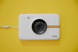 A white instant camera with a minimalist design features a lens in the center. A colorful stripe pattern in cyan, magenta, yellow, and orange runs horizontally on the camera front. A small flash is present on the upper left, and the word 'snap' is written on the bottom right. The camera rests on a bright yellow background, complemented by a white wrist strap attached to its side.