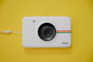 A white instant camera with a minimalist design features a lens in the center. A colorful stripe pattern in cyan, magenta, yellow, and orange runs horizontally on the camera front. A small flash is present on the upper left, and the word 'snap' is written on the bottom right. The camera rests on a bright yellow background, complemented by a white wrist strap attached to its side.