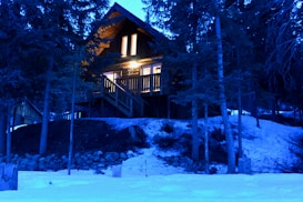 A cozy log cabin with warm glowing lights is nestled among tall evergreen trees. The surrounding area is blanketed in snow, creating a serene and peaceful winter scene. The cabin appears inviting against the backdrop of the evening sky.