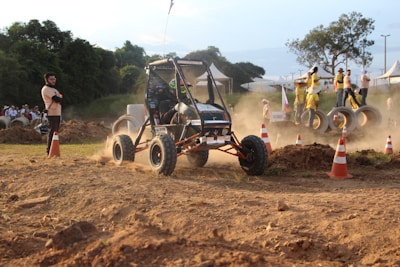 A small off-road vehicle is navigating a dusty terrain surrounded by traffic cones. The scene is set outdoors with spectators and officials standing nearby, some on large concrete pipes. The background includes greenery and temporary structures like tents.
