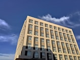 A modern multi-story building with large windows, set against a bright blue sky. White clouds are scattered across the sky, and a small airplane is visible in the distance to the left of the building.