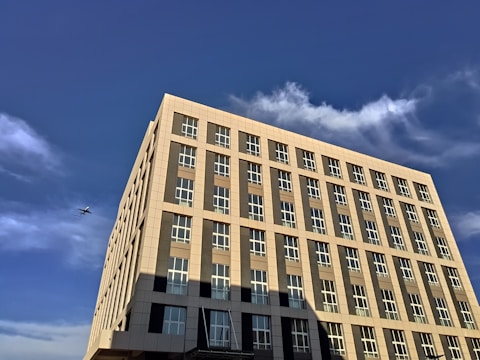 A modern multi-story building with large windows, set against a bright blue sky. White clouds are scattered across the sky, and a small airplane is visible in the distance to the left of the building.