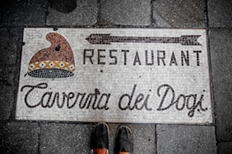 A mosaic tile sign embedded in a sidewalk reads 'Restaurant Taverna dei Dogi' with an illustration of a traditional hat. The tiles form a rustic and classic design, reflecting a vintage aesthetic.
