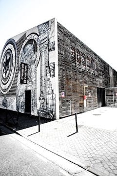A modern building featuring intricate black and white street art on its facade. The artwork includes abstract designs and large circular patterns. The structure is on a paved street and appears to be a public facility with signs near the entrance.