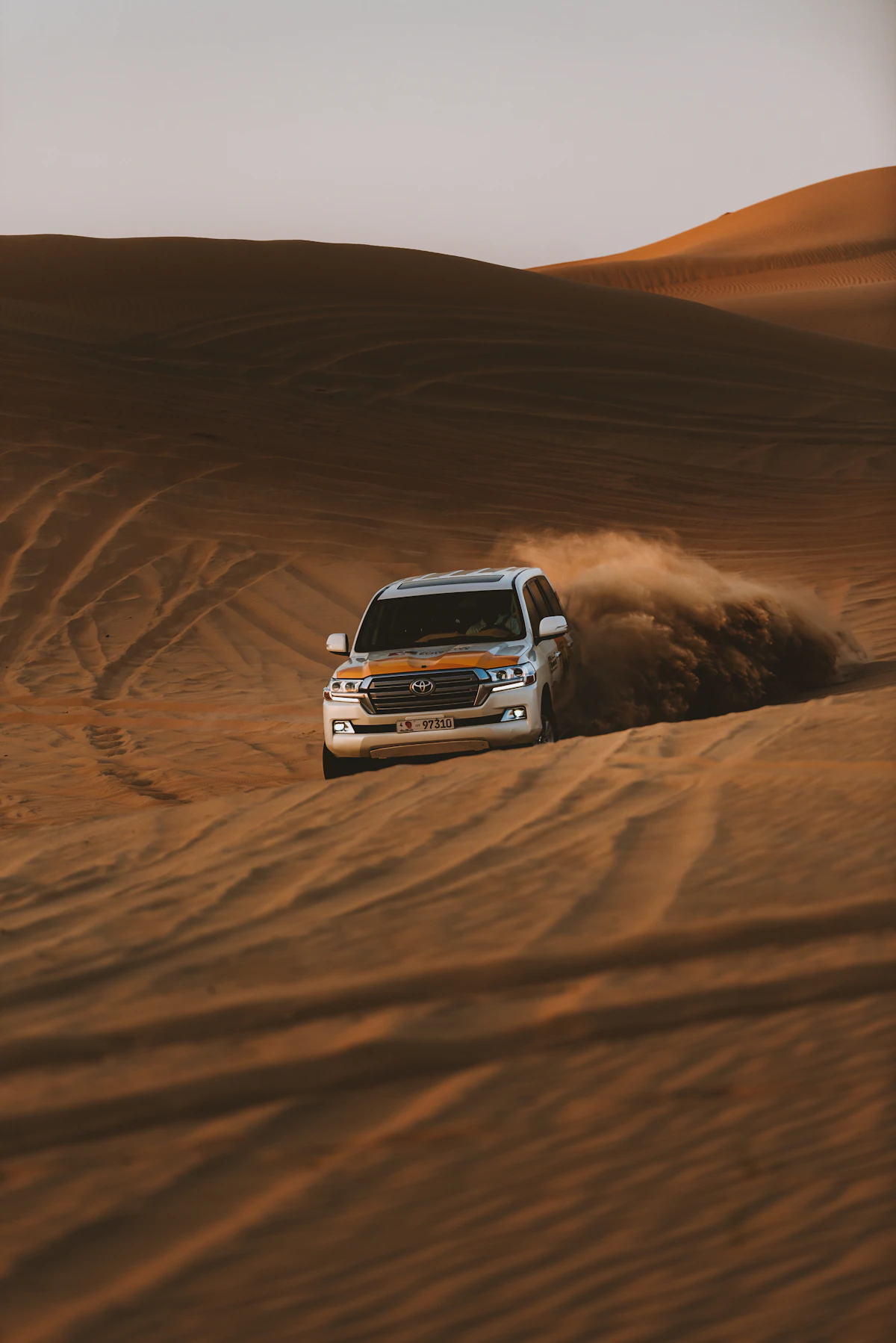A white SUV driving through sand dunes, kicking up a large cloud of dust