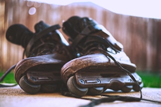 A close-up perspective of a pair of inline skates on a concrete surface, with laces loosely untied. The focus is on the worn wheels and scuffed material, indicating use. The background is softly blurred, suggesting an outdoor setting.