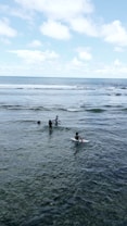Several people are in shallow ocean waters, engaging in activities like wading and possibly preparing to surf. The sky is predominantly clear with scattered clouds, and the sea is calm with a few small waves.