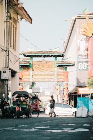 A bustling urban street scene features a large, ornately designed traditional Chinese gate in the background. Pedicabs and a motorcycle are parked along the street, and a man is casually walking nearby. Posters and advertisements are visible on the buildings, and colorful decorations adorn the scene.