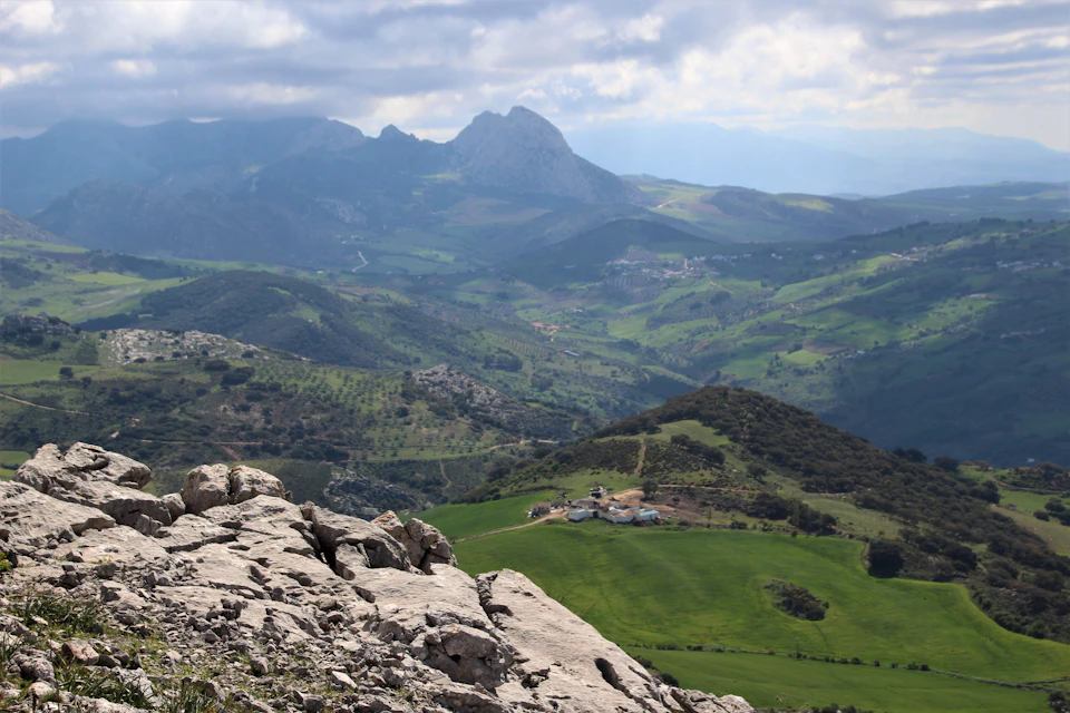 The image depicts a picturesque mountain landscape with a large, lush green valley below. The mountains are covered in trees and shrubs, creating a dense forested area on one side of the valley. In the center of the valley is a small village nestled among the greenery. The sky above is filled with clouds, casting a soft light over the scene.