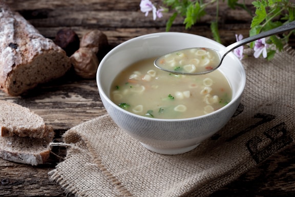 A rustic setting featuring a bowl of soup with small pasta and vegetables. A metal spoon is held above the bowl, resting on a burlap sack laid on a wooden table. Slices of crusty bread and whole bread are placed in the background alongside a few herbs and flowers.