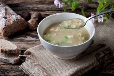 A rustic setting featuring a bowl of soup with small pasta and vegetables. A metal spoon is held above the bowl, resting on a burlap sack laid on a wooden table. Slices of crusty bread and whole bread are placed in the background alongside a few herbs and flowers.