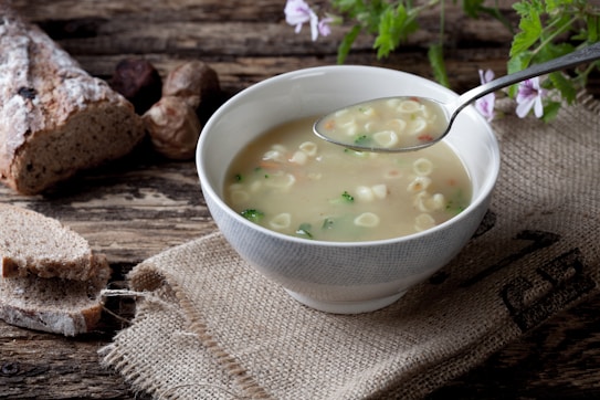 A rustic setting featuring a bowl of soup with small pasta and vegetables. A metal spoon is held above the bowl, resting on a burlap sack laid on a wooden table. Slices of crusty bread and whole bread are placed in the background alongside a few herbs and flowers.
