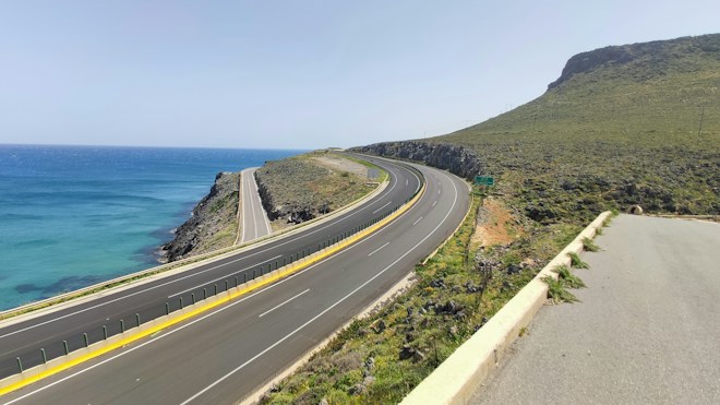 A coastal highway stretches alongside a vast expanse of blue ocean. The road curves gently around a grassy hill. The scene is calm and clear, with minimal traffic and a bright sky.