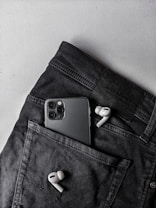 A pair of white wireless earbuds and a smartphone with multiple cameras are placed on a dark pair of pants. The smartphone is partially tucked into the pocket, while the earbuds are positioned outside.
