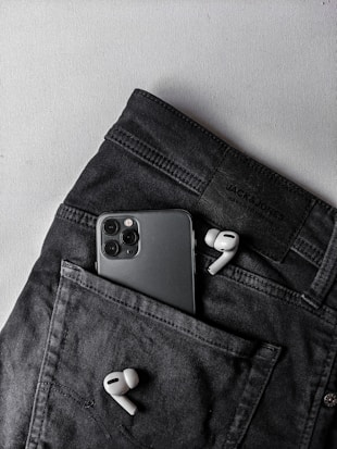 A pair of white wireless earbuds and a smartphone with multiple cameras are placed on a dark pair of pants. The smartphone is partially tucked into the pocket, while the earbuds are positioned outside.