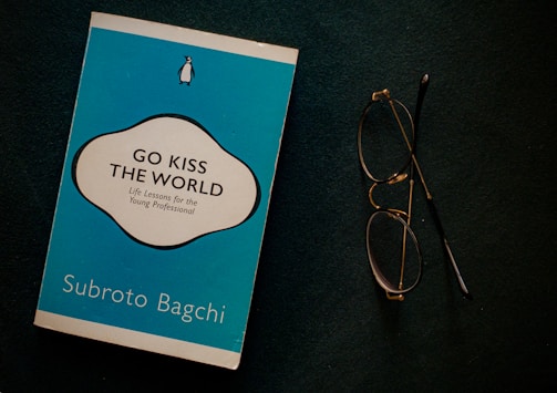 A blue book titled 'Go Kiss the World' by Subroto Bagchi is placed next to a pair of round eyeglasses on a dark surface. The book cover features a white and black circular design with text and a small penguin logo at the top.