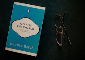 A blue book titled 'Go Kiss the World' by Subroto Bagchi is placed next to a pair of round eyeglasses on a dark surface. The book cover features a white and black circular design with text and a small penguin logo at the top.