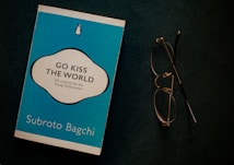 A blue book titled 'Go Kiss the World' by Subroto Bagchi is placed next to a pair of round eyeglasses on a dark surface. The book cover features a white and black circular design with text and a small penguin logo at the top.
