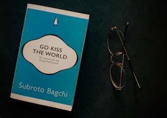 A blue book titled 'Go Kiss the World' by Subroto Bagchi is placed next to a pair of round eyeglasses on a dark surface. The book cover features a white and black circular design with text and a small penguin logo at the top.