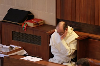 An elderly person dressed in a white religious robe is seated in a wooden pew with their head resting on their hand, possibly deep in thought or prayer. A red book with colorful ribbons is placed beside them. The setting appears to be a quiet and solemn environment, possibly a church or chapel.