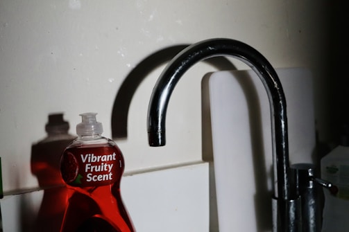 A kitchen sink with a metallic faucet stands beside a red dishwashing liquid bottle labeled 'Vibrant Fruity Scent.' The lighting casts shadows on the wall, adding contrast to the scene.