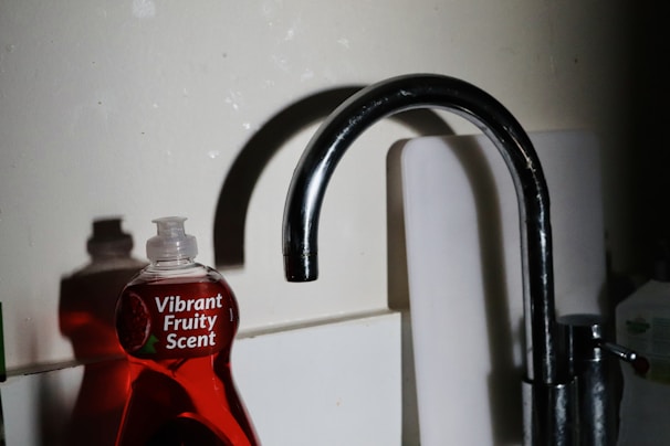 A kitchen sink with a metallic faucet stands beside a red dishwashing liquid bottle labeled 'Vibrant Fruity Scent.' The lighting casts shadows on the wall, adding contrast to the scene.