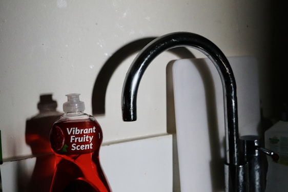 A kitchen sink with a metallic faucet stands beside a red dishwashing liquid bottle labeled 'Vibrant Fruity Scent.' The lighting casts shadows on the wall, adding contrast to the scene.