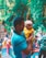 A man holds a young child in his arms, surrounded by a lush, green outdoor setting. The child is wearing a yellow outfit with red bangles and is being gently supported. The background is vibrant, with out-of-focus plants and a colorful staircase, suggesting a lively, possibly festive environment.