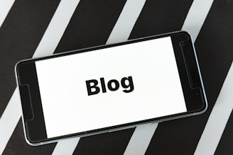 A smartphone displaying the word 'Blog' on its screen is placed on a surface with alternating black and white diagonal stripes. The phone's screen is easily readable and prominently features the text in bold.