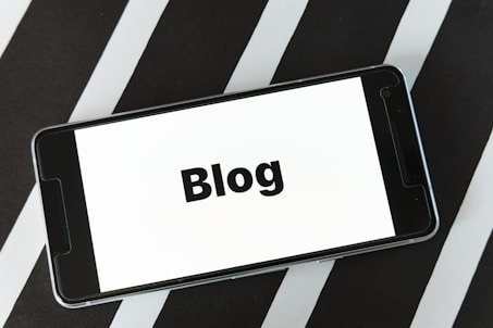 A smartphone displaying the word 'Blog' on its screen is placed on a surface with alternating black and white diagonal stripes. The phone's screen is easily readable and prominently features the text in bold.