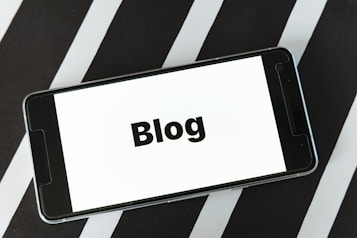 A smartphone displaying the word 'Blog' on its screen is placed on a surface with alternating black and white diagonal stripes. The phone's screen is easily readable and prominently features the text in bold.