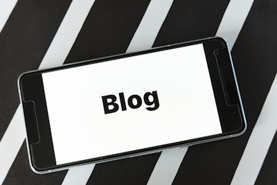 A smartphone displaying the word 'Blog' on its screen is placed on a surface with alternating black and white diagonal stripes. The phone's screen is easily readable and prominently features the text in bold.