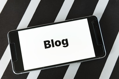 A smartphone displaying the word 'Blog' on its screen is placed on a surface with alternating black and white diagonal stripes. The phone's screen is easily readable and prominently features the text in bold.