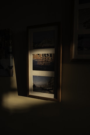 A framed piece containing multiple photographs hangs on a wall. Each photograph is individually bordered within the frame, and varying levels of light and shadow play across its surface.