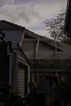 A partially visible rooftop of a building with a triangular structure. There is a modern design with overlapping wooden panels, a corrugated metal roof, and nearby vegetation. In the foreground, there is a wooden fence and foliage, and the background shows a cloudy sky.