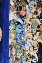 A vibrant display of ornate and intricately designed Venetian masks. The collection features masks with various patterns, embellishments, and colors, including red, gold, blue, and white. Some masks have elaborate designs with tassels and beading, while others are painted with swirls and geometric patterns. The masks are arranged closely together, and some feature theatrical and festive motifs.
