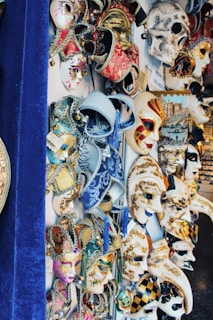 A vibrant display of ornate and intricately designed Venetian masks. The collection features masks with various patterns, embellishments, and colors, including red, gold, blue, and white. Some masks have elaborate designs with tassels and beading, while others are painted with swirls and geometric patterns. The masks are arranged closely together, and some feature theatrical and festive motifs.