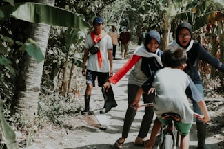 A group of people are enjoying a walk on a dirt path surrounded by lush banana trees and greenery. Two women wearing headscarves interact playfully with a child on a bicycle. A man walks behind them, and two more people are visible further down the path. The scene suggests casual outdoor activity and camaraderie.