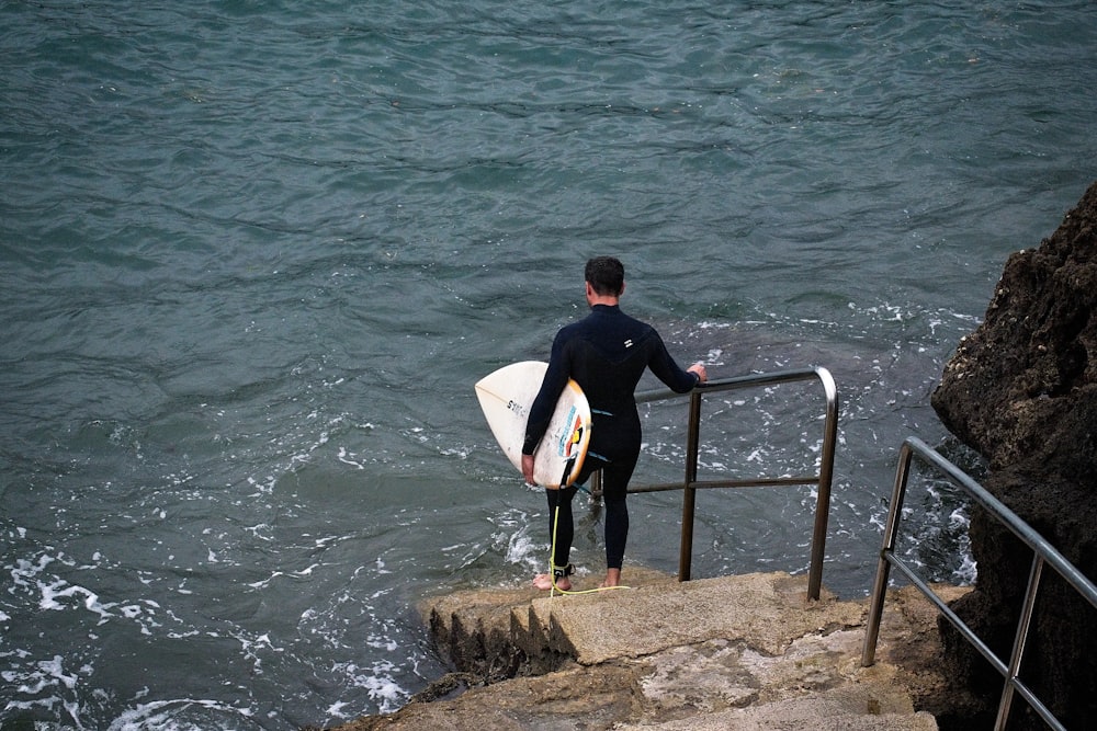 水域の近くの茶色の岩の上に立って白いサーフボードを運ぶ黒いシャツの男