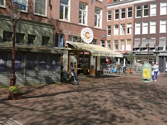 A street scene featuring a red-brick building housing a shop labeled 'Bagels & Beans' with a cream-colored awning. In front of the shop, there is a row of bicycles parked on the sidewalk. A person in a blue outfit is walking with a bag. A vintage-style lamppost is situated on the left, and shadows of trees are cast on the ground.